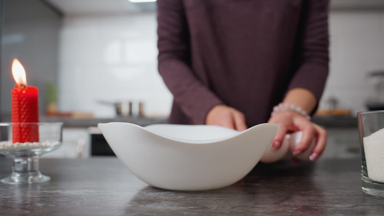 Visuale parziale di una donna che si volta con una ciotola bianca in mano, appoggiandola delicatamente sul tavolo mentre sistema le uova, mano adornata da un braccialetto, candela rossa accesa sul tavolo, cucina sullo sfondo