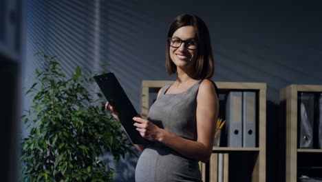 pregnant businesswoman in glasses reading documents in the office and then smiles at the camera