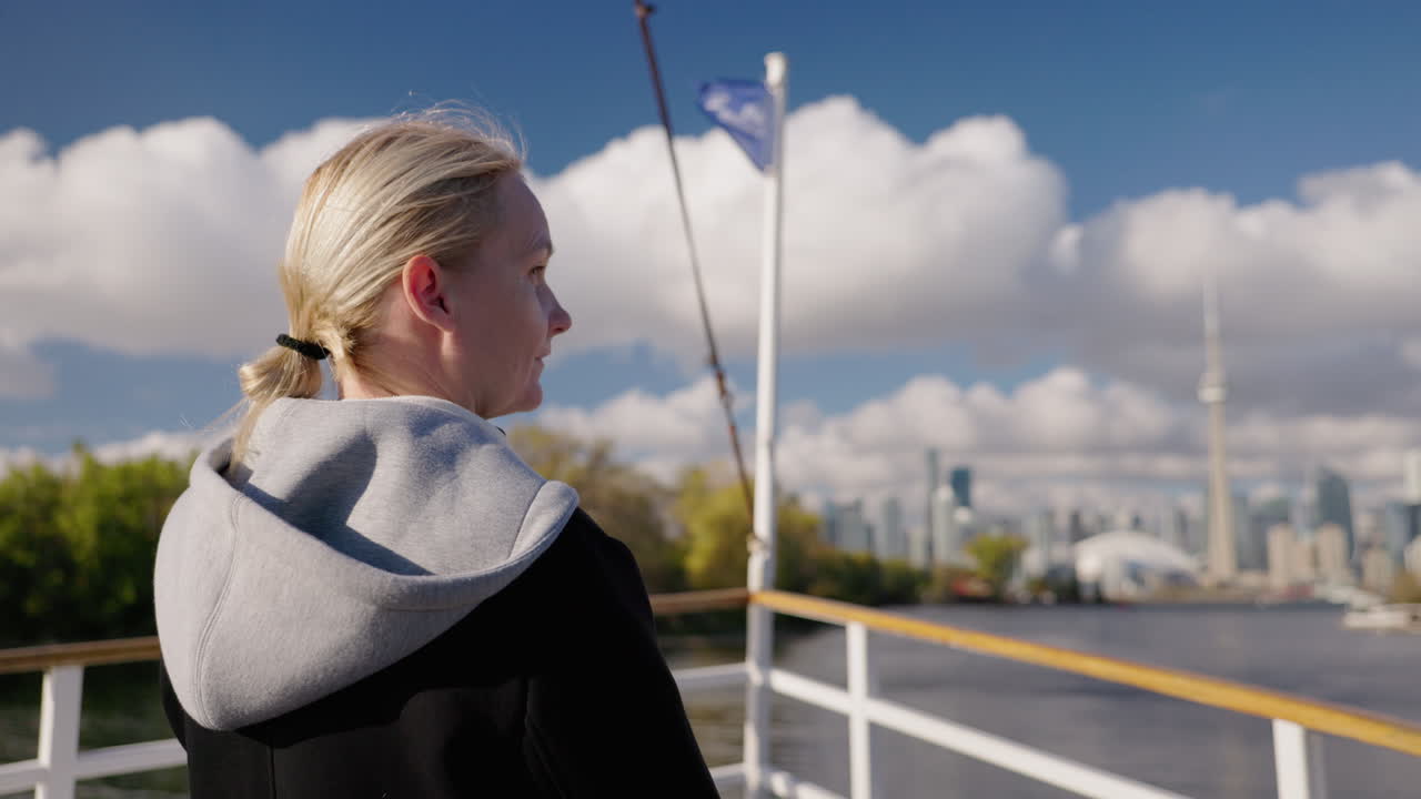 Giovane donna turista, di spalle, con i capelli biondi, sul ponte di una barca, osserva lo skyline di Toronto attraverso il Lago Ontario, in un'atmosfera di viaggio serena sul lungolago.
