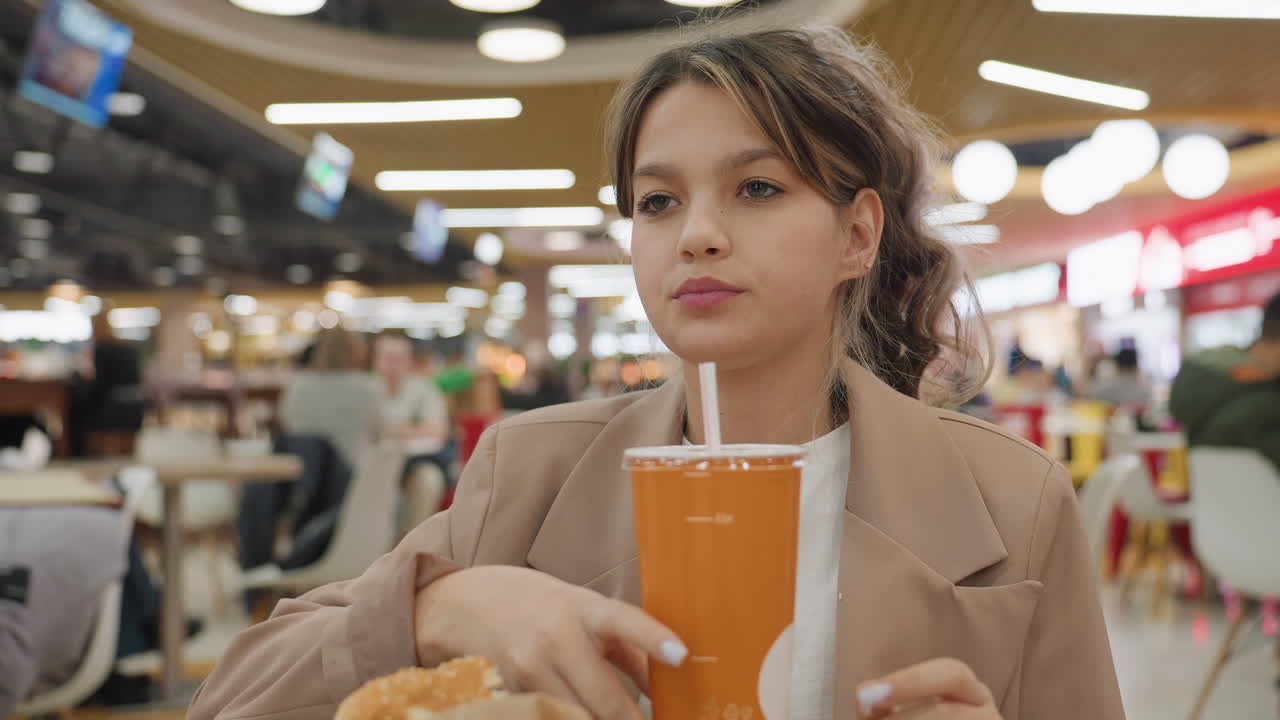 Adolescente observando pensativamente su entorno; joven caucásico contemplando en silencio una concurrida zona de comida; joven adolescente caucásico que parece reflexivo mientras observa una escena distante en un concurrido patio de comidas.