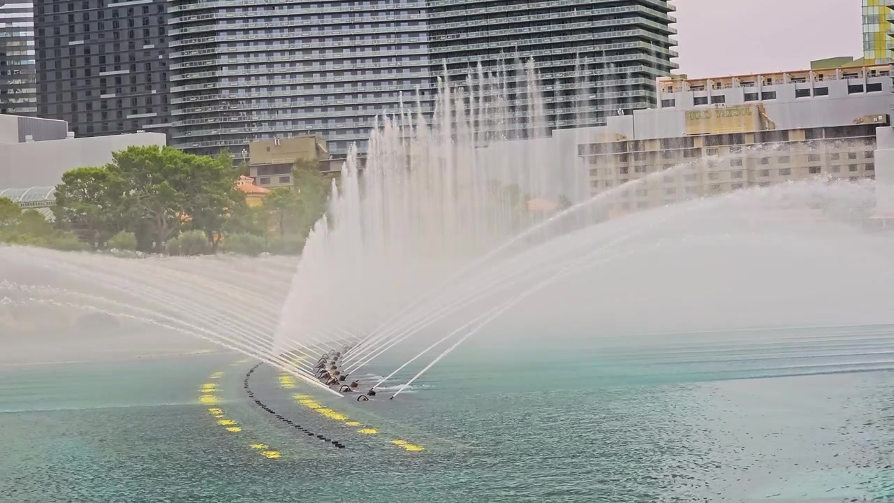 Espectáculo diurno de fuentes en un resort con hileras de chorros de agua sincronizados, suave neblina, superficie de lago turquesa, modernas torres de hotel, arcos de piedra y un soleado paisaje urbano de viaje para el turismo