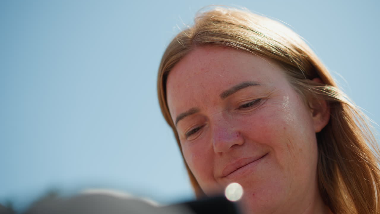 Close up of digital creator smiling warmly while going through phone under clear blue sky, soft sunlight on face, peaceful outdoor mood showing creativity