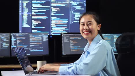 side view of asian female programmer smiling to camera while writing code by a laptop using multiple monitors showing database on terminal window desktops in the office