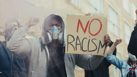 vista de cerca del hombre afroamericano con máscara de gas sosteniendo carteles contra el racismo y manifestándose con otras personas en la calle