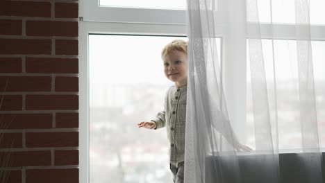 boy with blonde hair hiding behind curtain at home, waving out there