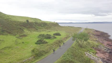 three people cycling on a bike over a black asphalt roat between the green meadows and lovely view at great cumbrae, scotland on a cloudy day