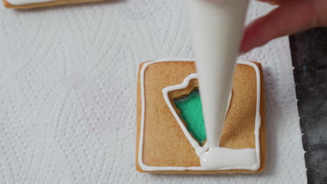 close-up of person icing a cookie with white frosting, creating intricate designs around a green filling, on textured paper, showcasing the baking and decoration process