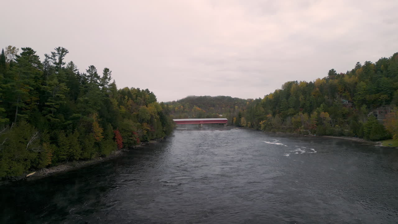 A drone view approaching a red covered bridge over a flowing river in autumn