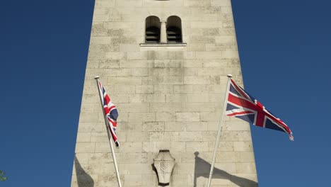 Torre-Del-Reloj-Conmemorativo-De-La-Guerra-En-Inglaterra,-Con-Banderas-Británicas-Moviéndose-En-El-Viento,-En-4k,-Cámara-Lenta