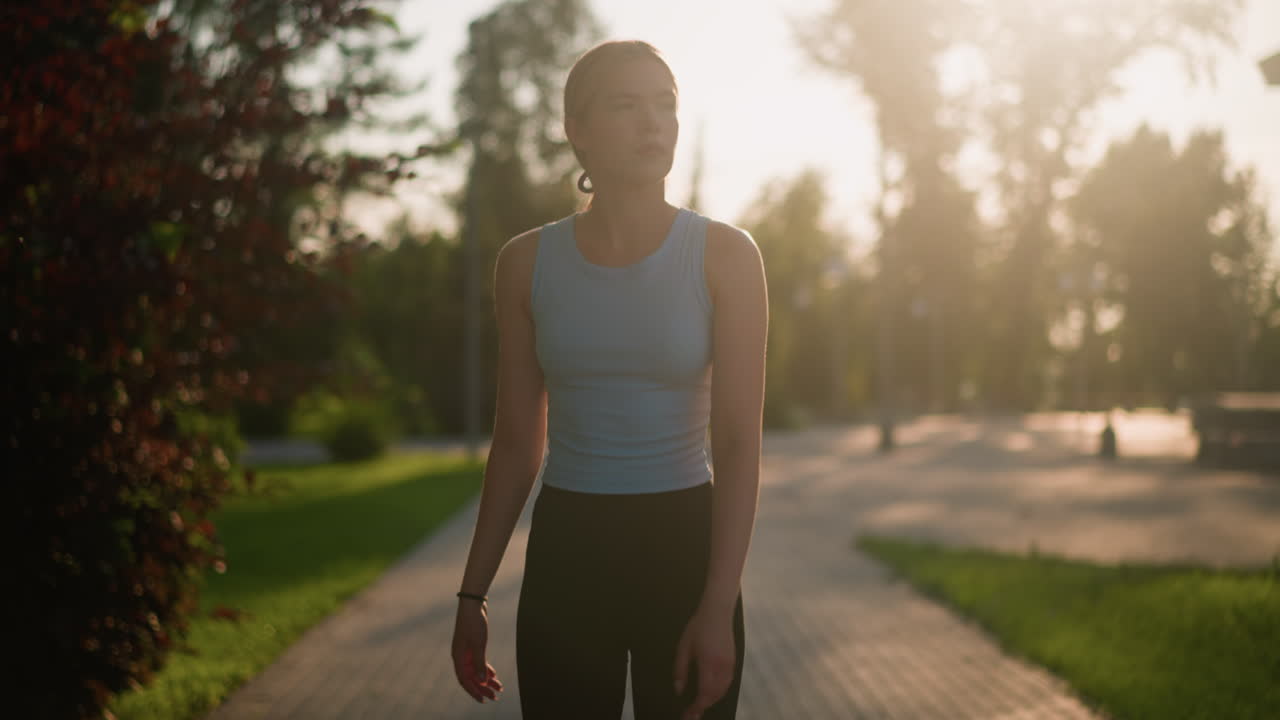 Young lady looking thoughtful while roller skating outdoors on sunny day, swinging her hands for balance, sunlight creating a glowing effect around her, blurred background of lush greenery, trees