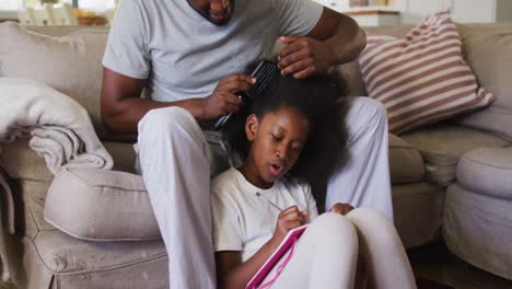 Afroamerikanischer-Vater-Bürstet-Die-Haare-Seiner-Tochter,-Während-Er-Zu-Hause-Auf-Der-Couch-Sitzt