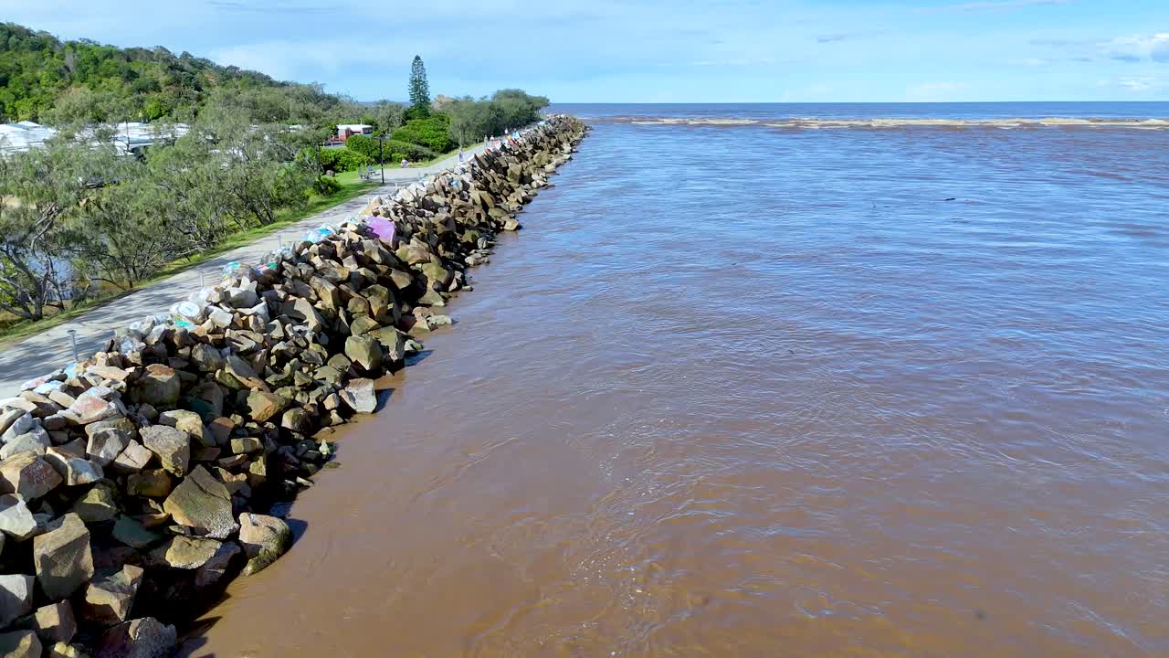 Drone camera moves steadily above a stone breakwater along a brown river, bordered by greenery, under bright daylight in Nambucca, NSW, Australia