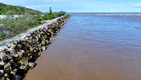 drone camera moves steadily above a stone breakwater along a brown river, bordered by greenery, under bright daylight in nambucca, nsw, australia