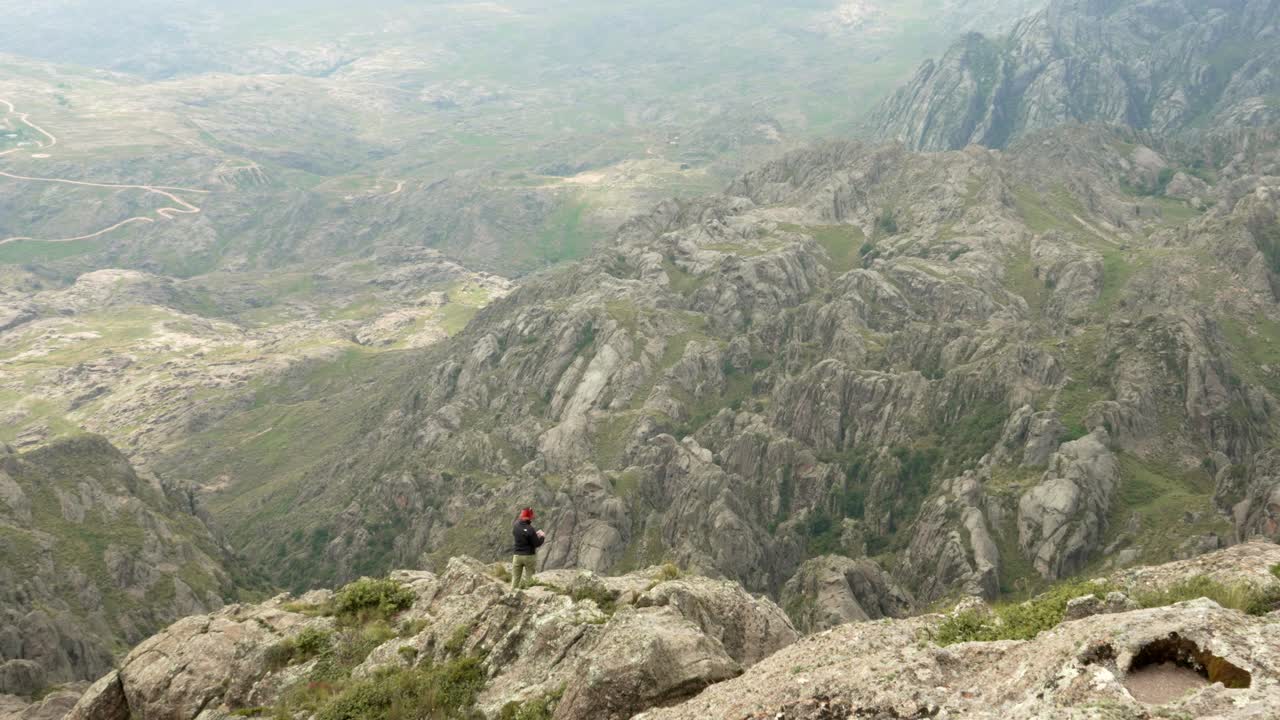 Handheld shot of a woman hiker standing on a rocky cliff overlooking the vast mountain landscape of Los Gigantes, Córdoba, Argentina