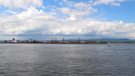 wide view of sailboat far away sailing along coast port with calm water and cloudy sky in horizon, quebec region