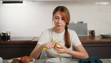 portrait view of woman in apron peeling raw potatoes with peeler over bowl on kitchen table, surrounded by dishes and utensils in clean modern home cooking environment