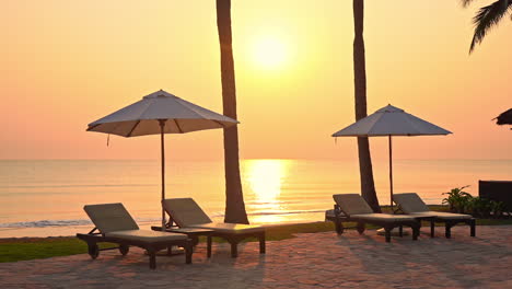 Lovely-view-of-a-golden-sunset-with-in-some-tropical-destination,-with-empty-beach-lounges-and-umbrellas-in-the-foreground-and-clear-orange-sky-and-golden-sun-reflecting-on-the-surface-of-ocean