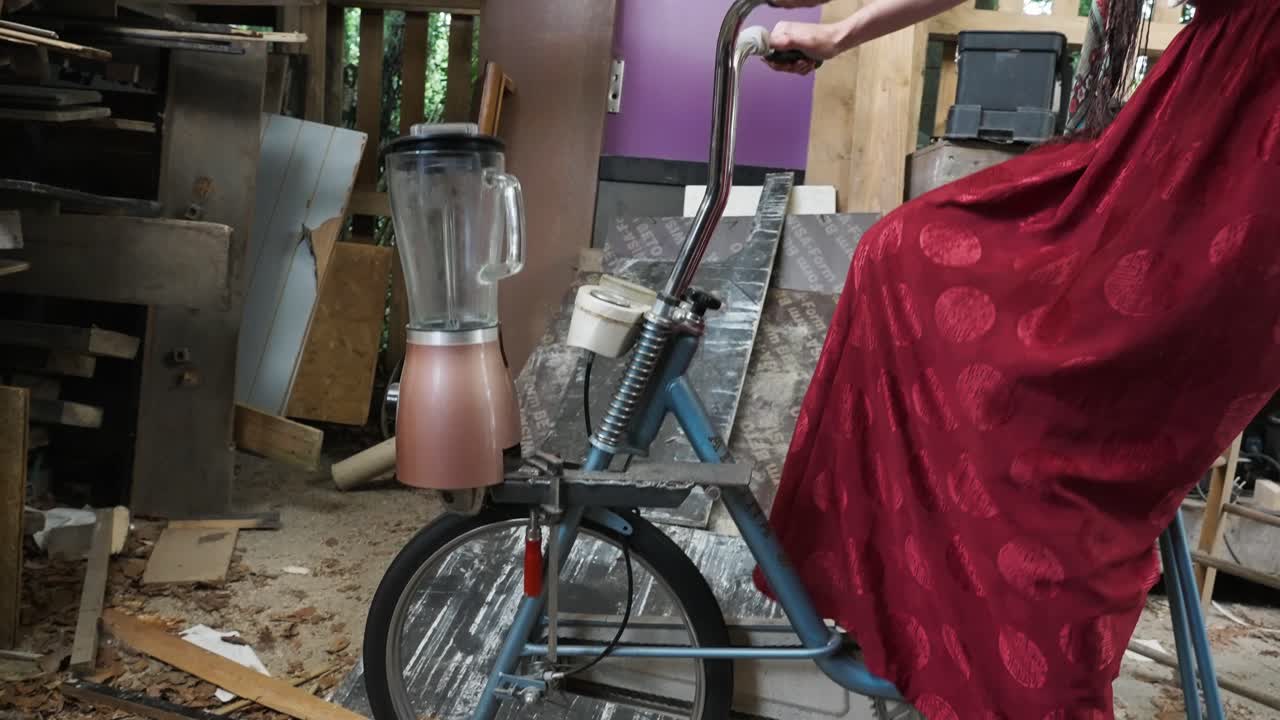 sonriente hermosa mujer eco consciente hippie pedalingope en bicicleta reutilizada reciclada ejecutando una licuadora alimentada por humanos dentro del cobertizo