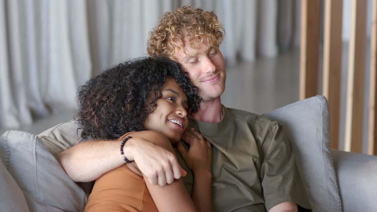 Diverse couple sitting on gray sofa at home, man gesturing playfully, woman leaning in for comfort
