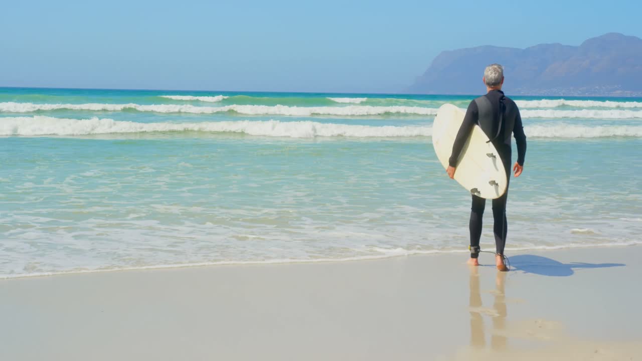 vista trasera de un surfista masculino caucásico senior activo caminando con una tabla de surf en la playa 4k