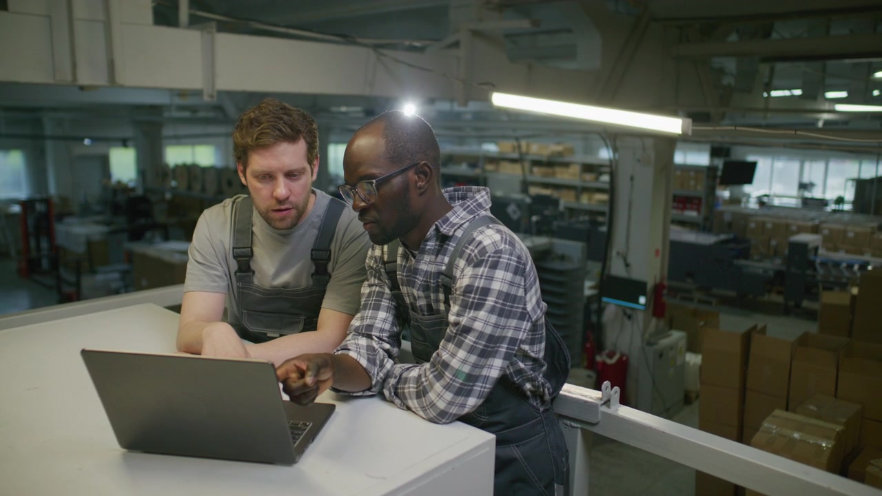 Multiethnic Engineers Discussing Data on Laptop in Printing Center Warehouse