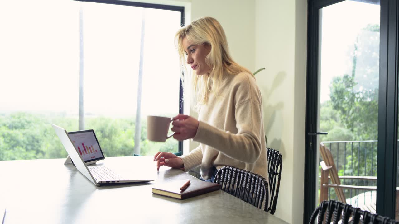 Lavoratrice da remoto che nota un grafico a barre sul laptop mentre analizza e sorseggia un caffè prendendo appunti a casa
