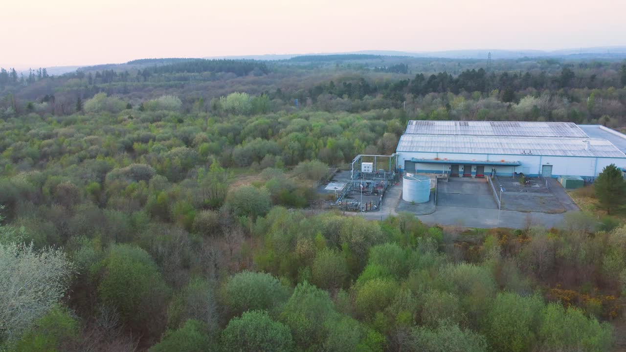 Sideways Aerial View Over Run Down Warehouse with Loading Bays Surrounded by Forest Landscape. Industrial Infrastructre Surrounded by Natural Environment. 4K