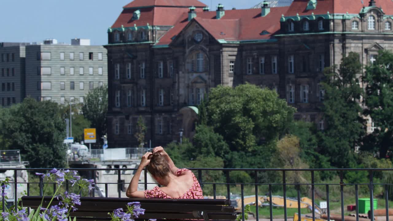 Woman sits on bench by riverbank, enjoying sunny day and cityscape with historic building backdrop