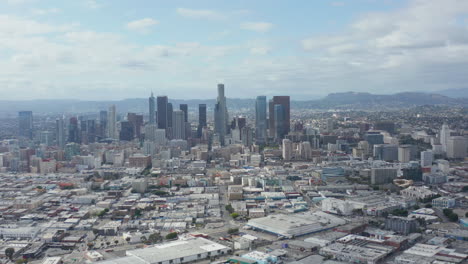 AERIAL:-Slow-Side-Shot-of-Downtown-Los-Angeles-Skyline-with-Warehouse-Art-District-in-Foreground-with-Blue-Sky-and-Clouds