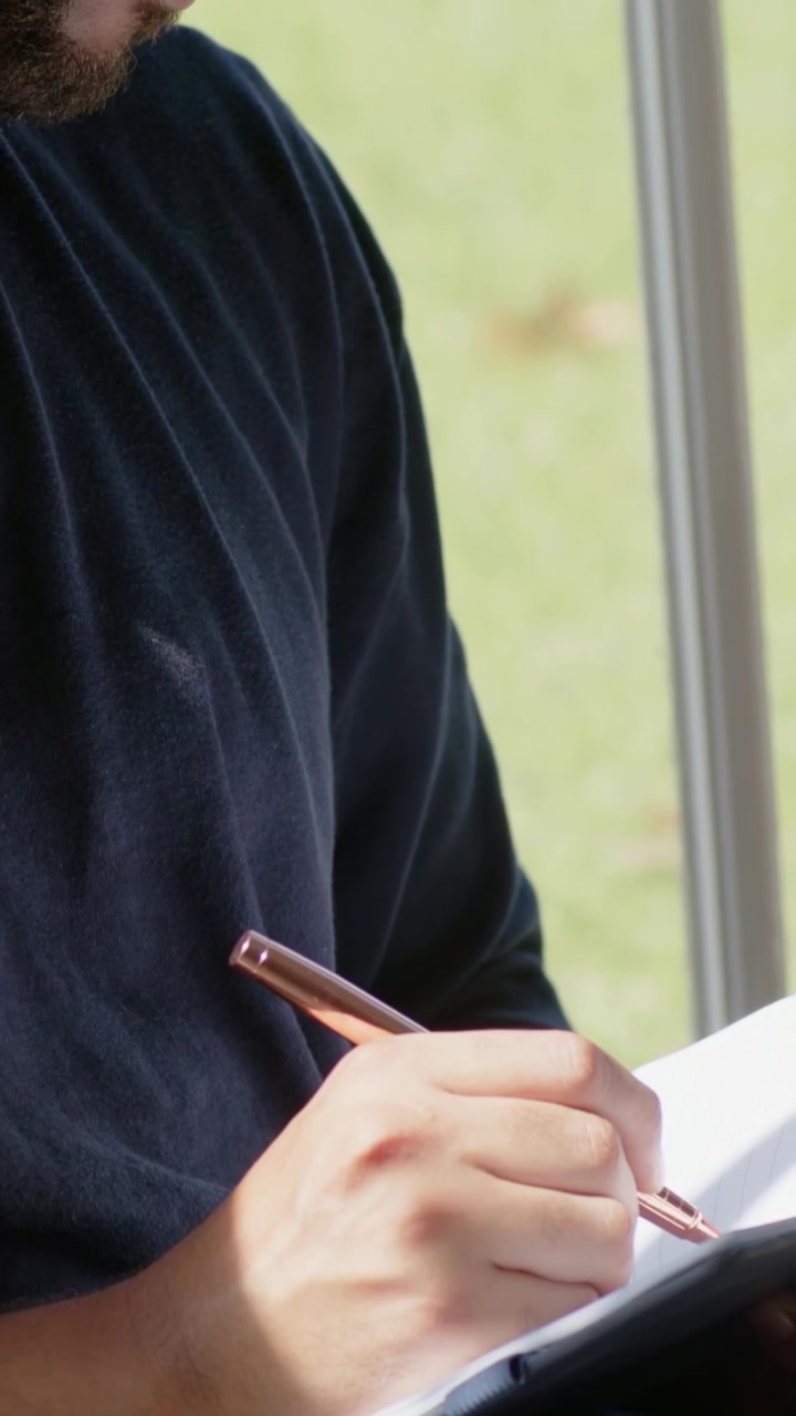 Vertical video: Looking down, man in dark shirt noting at window with pen and notebook, copy space