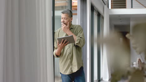 thoughtful senior biracial man standing by window using tablet at home, copy space, slow motion