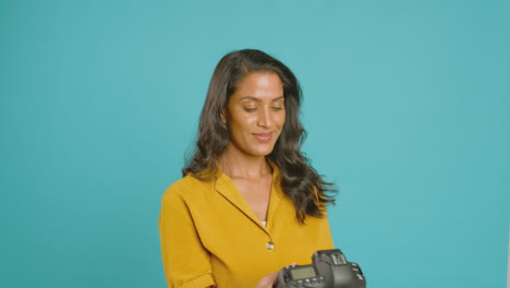 Mature-Female-Photographer-With-Camera-Against-Blue-Background-On-Shoot-In-Studio