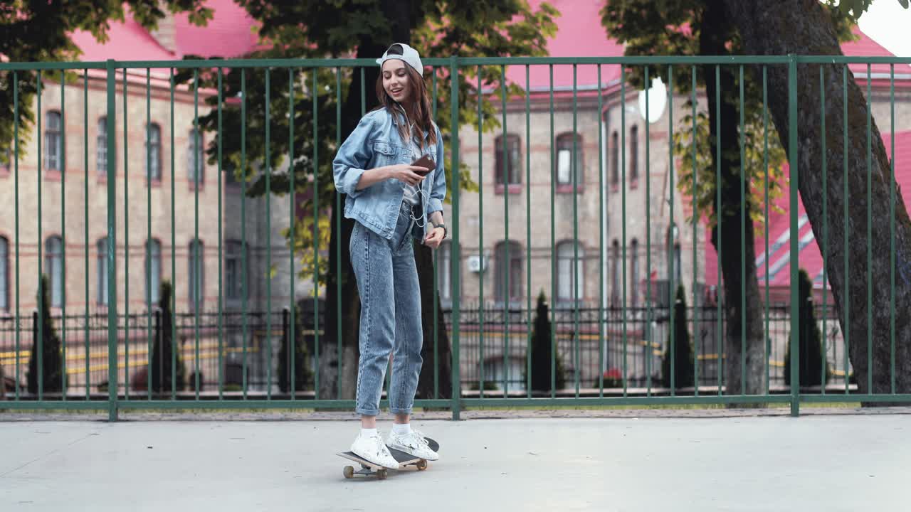 Full length view of a teenage girl listening music with smartphone while standing on her skateboard and dancing in the street