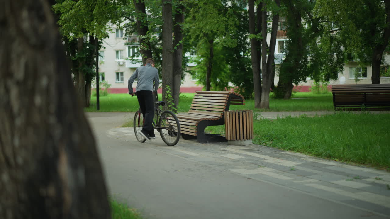 회색 을 입은 어린 소년은 조용한 나무로 둘러싸인 공원을 자전거로 타고, 나무 벤치에 멈추고, 자전거를 주차하고, 울창한 초록색으로 둘러싸여 도시 환경에서 자연을 즐기며 앉습니다.