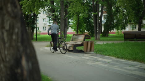 young boy in gray top rides bicycle through serene tree-lined park, stops by wooden bench, parks bike, and sits down, surrounded by lush greenery enjoying nature in an urban environment