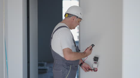 un hombre electricista con casco en el apartamento comprueba el trabajo de los enchufes y interruptores después de la reparación