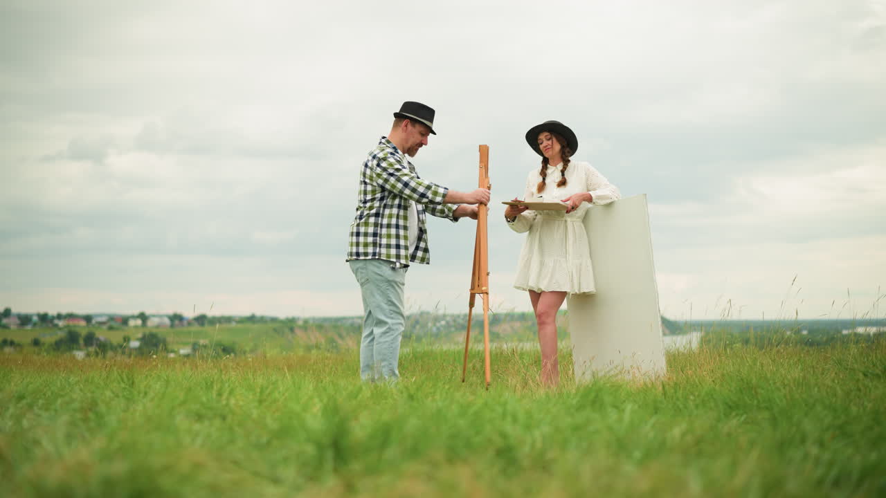 한 부부는 야외에서 그림을 그리기 위해 준비하는 동안 잔디에 서 있습니다. 색 셔츠와 검은 모자를 입은 남자는 색 드레스를 입은 여자가 스을 세우고 있습니다.