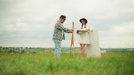 una pareja está de pie en un campo cubierto de hierba mientras se preparan para pintar al aire libre. el hombre, vestido con una camisa a cuadros y un sombrero negro, está montando un caballete, mientras que la mujer, vestida con un vestido blanco y