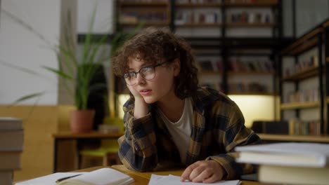 A-tired-brunette-girl-with-curly-hair-and-glasses-reads-documentation-at-a-table-in-the-library