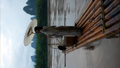 niña hanfu arreglando su cabello y sosteniendo una sombrilla mientras está de pie en una balsa de bambú junto al río li