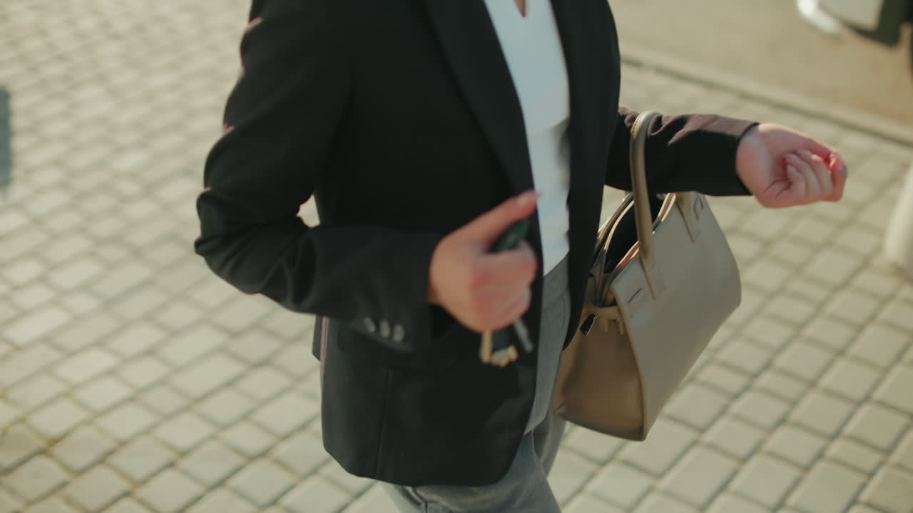 Partial view of woman retrieving key from handbag outdoors, people walking in background with parked cars nearby, professional attire visible, metal railings bordering walkway