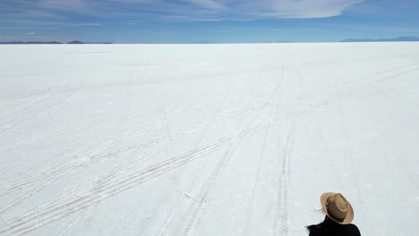 aerial overtakes woman walking toward flat horizon on uyuni salt flat