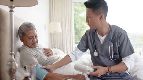 diverse male doctor testing blood pressure of senior male patient in bed at home, slow motion