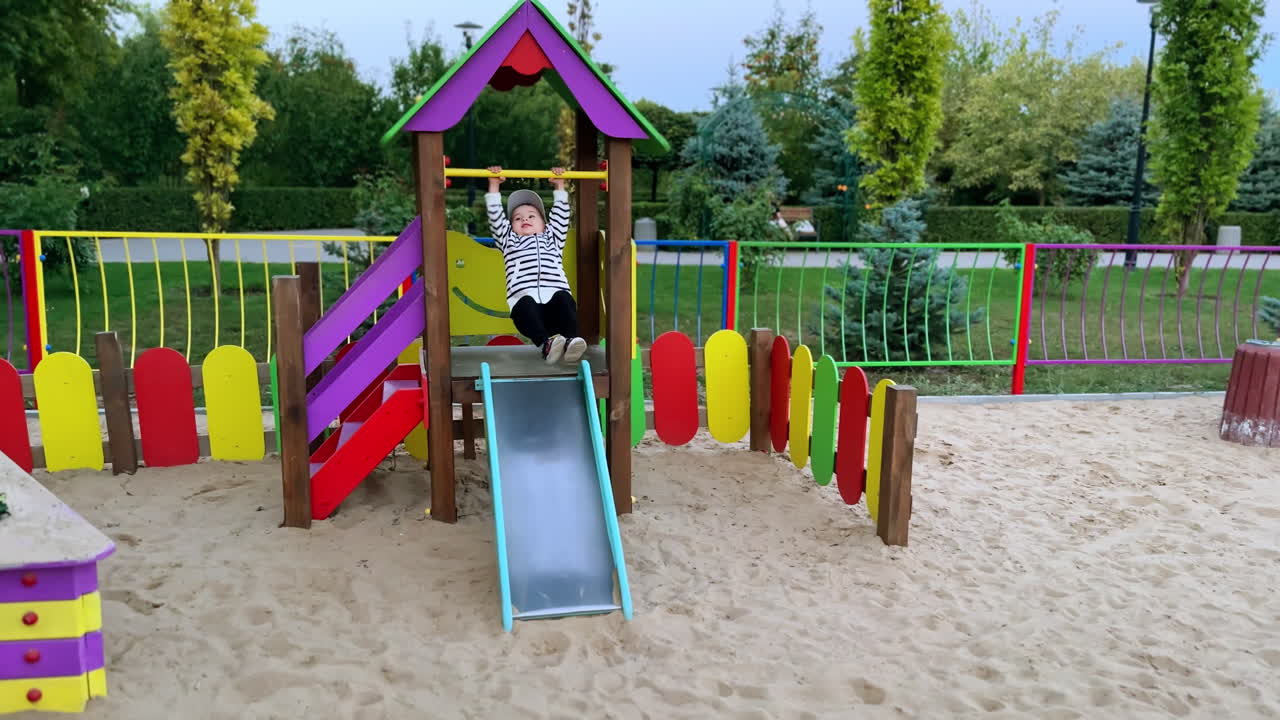 Cute baby boy having fun on the playground. Active kid hits his feet by the metal slide and smiles happily.