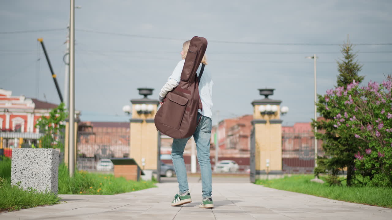female guitarist walking with guitar case through urban park pathway, pauses at wooden pillar to adjust strap, looks toward skyline under overcast sky, green grass and city architecture frame casual