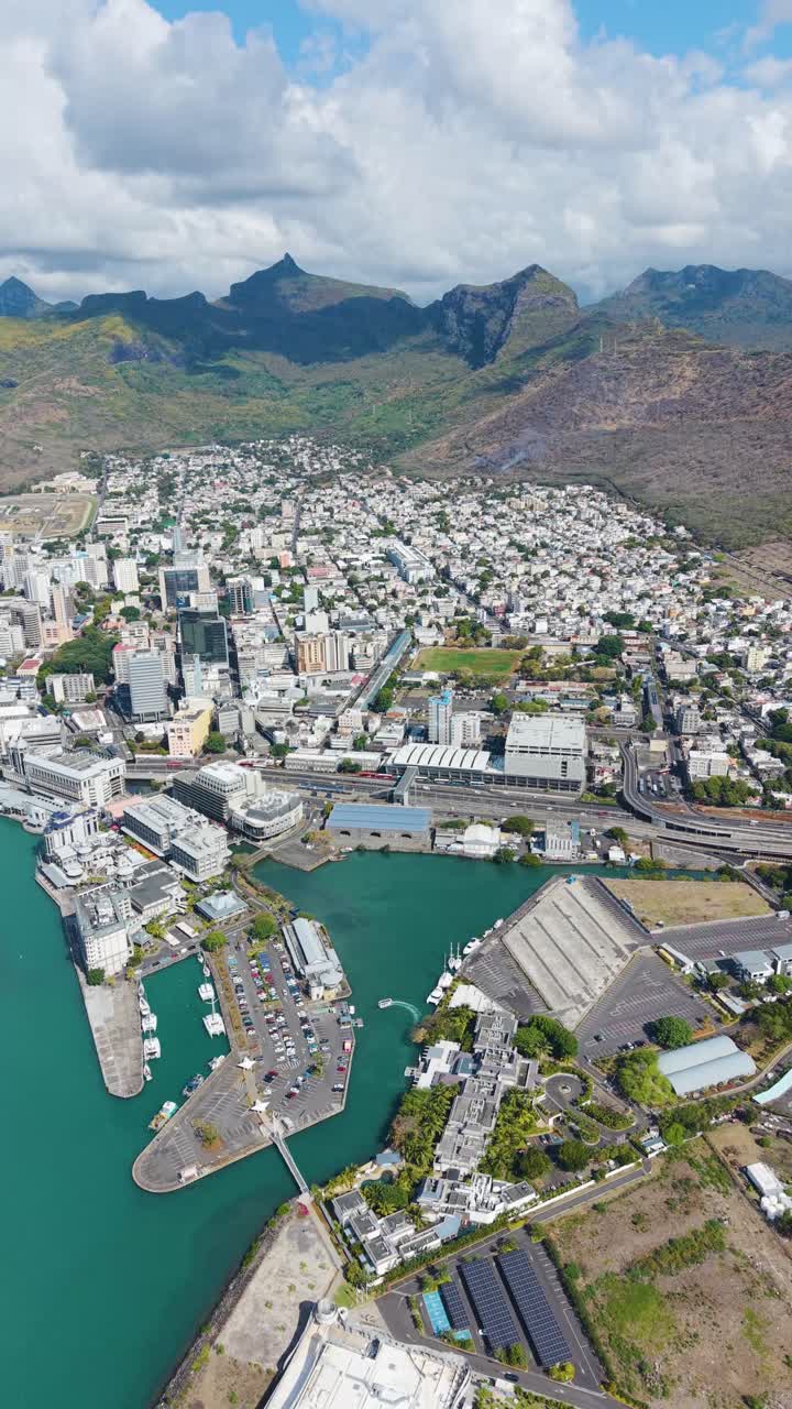 Vertical aerial view of Port Louis skyline in Mauritius. Features the bustling Caudan Waterfront, with the city nestled against the Moka Mountains and the blue ocean