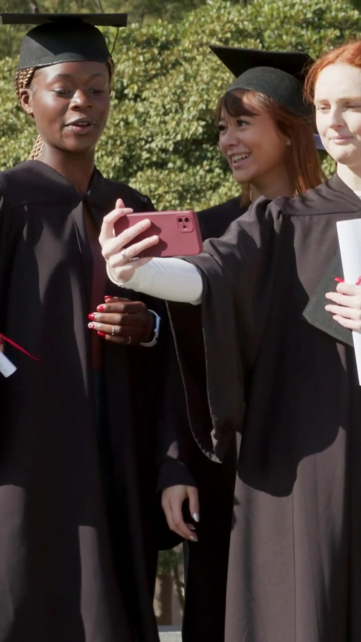 Video verticale: Una donna dai capelli rossi in abiti neri che si avvicina e offre il telefono per un selfie ad amici in posa sui gradini del campus.