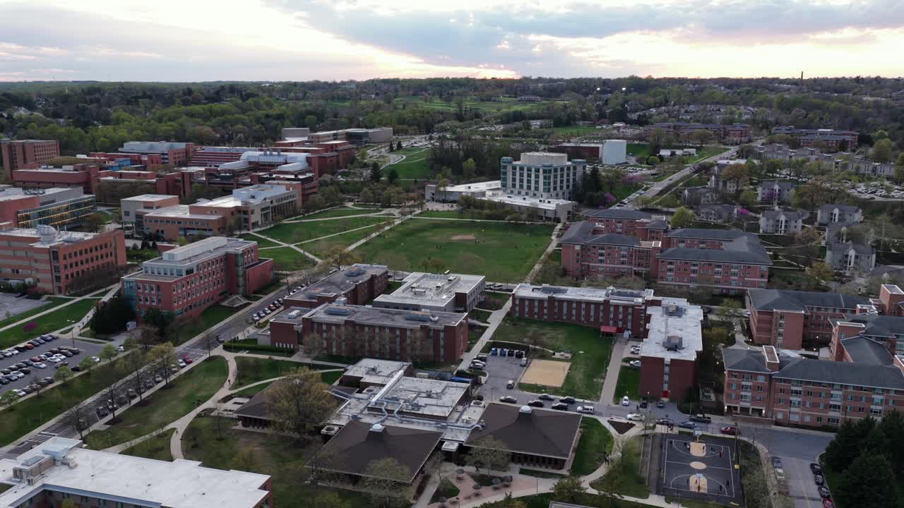 Ampia ripresa aerea del campus universitario del Maryland a Baltimora con edifici storici in mattoni, cortili verdi, sentieri e strutture accademiche sotto una soffusa luce serale in un contesto urbano.