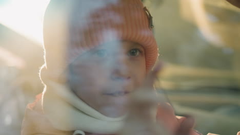 close-up of a young girl in a pink knit hat and scarf playing with her fingers, her eyes twinkling with curiosity as sunlight filters through the window