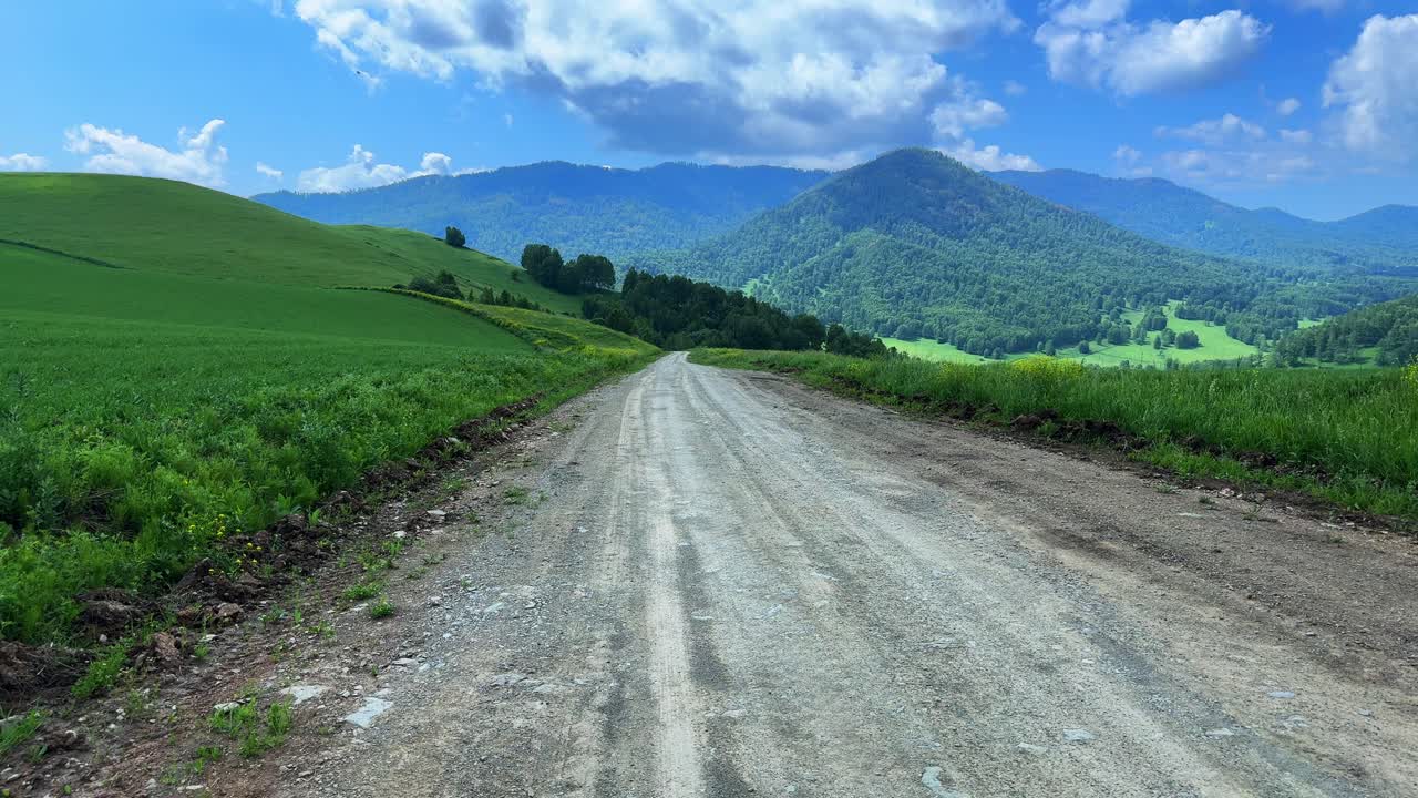 A Serene Dirt Road Through Lush Green Fields and Rolling Mountains Under a Bright Blue Sky with Fluffy White Clouds